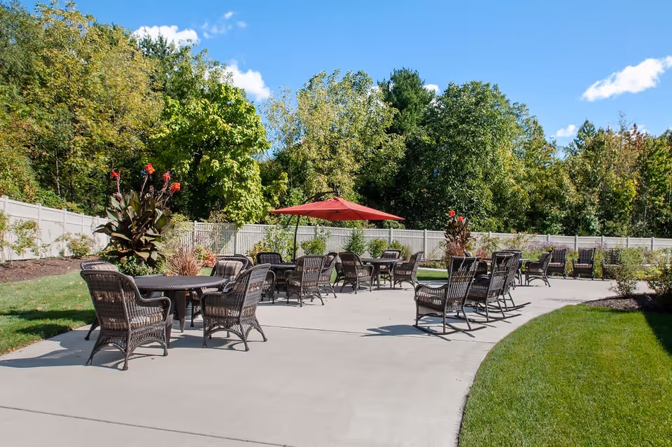 Outdoor patio with multiple wicker tables and chairs and a red umbrella on a paved area beside lawn and trees.