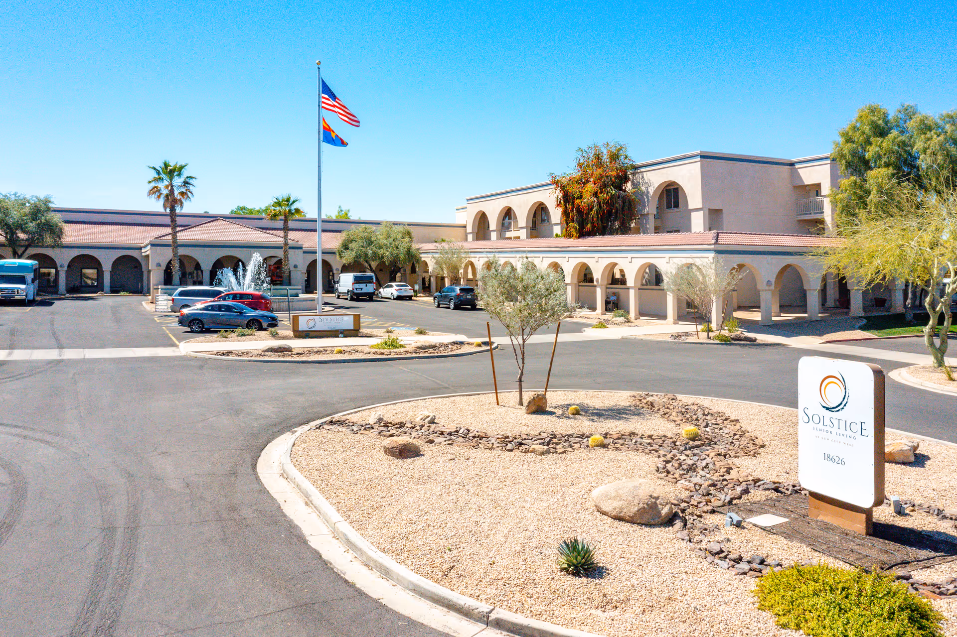 Front entrance of a beige Mediterranean-style senior living building with arched walkways, a fountain, flagpoles, parked cars and a Solstice sign.