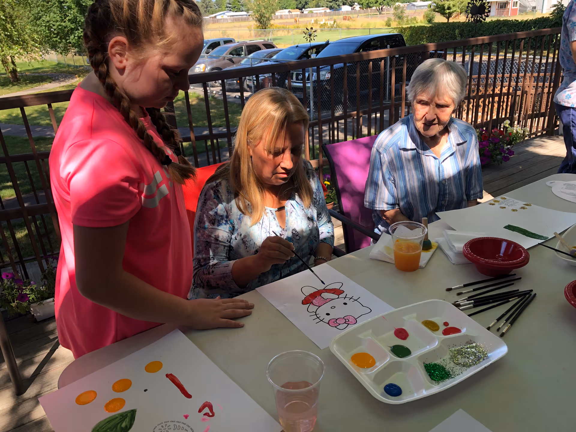 A caregiver assists elderly residents painting at an outdoor patio table.