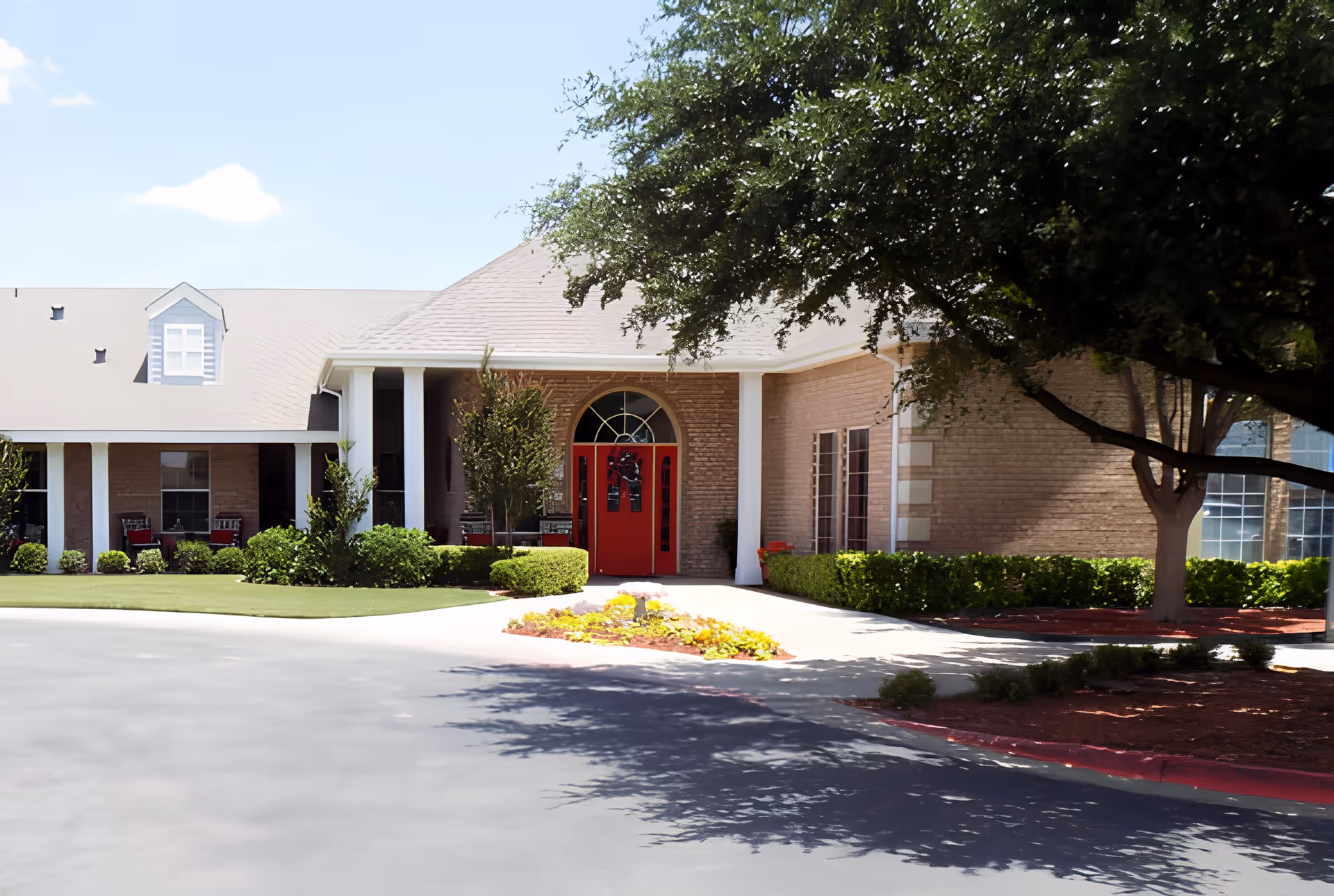 Front exterior view of a single-story brick building with a gray roof and a bright red double door entrance. The building is surrounded by neatly trimmed bushes, small trees, and a landscaped flower bed in front of the entrance. A large tree casts shadows on the driveway leading up to the entrance under a clear blue sky.
