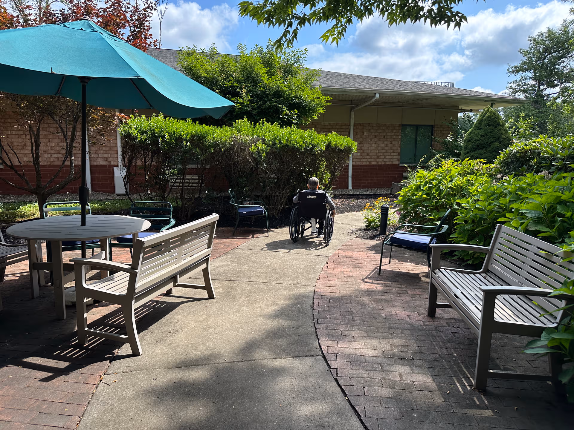 Outdoor courtyard at Complete Care at Park Place with benches, table, umbrella, and a wheelchair user.
