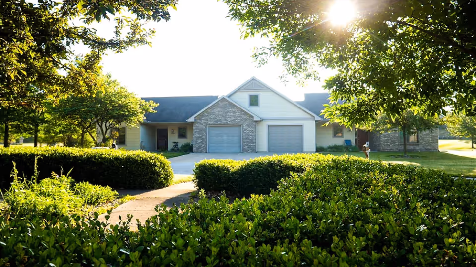 A single-story residential building with two garage doors, surrounded by green bushes and trees under bright sunlight.