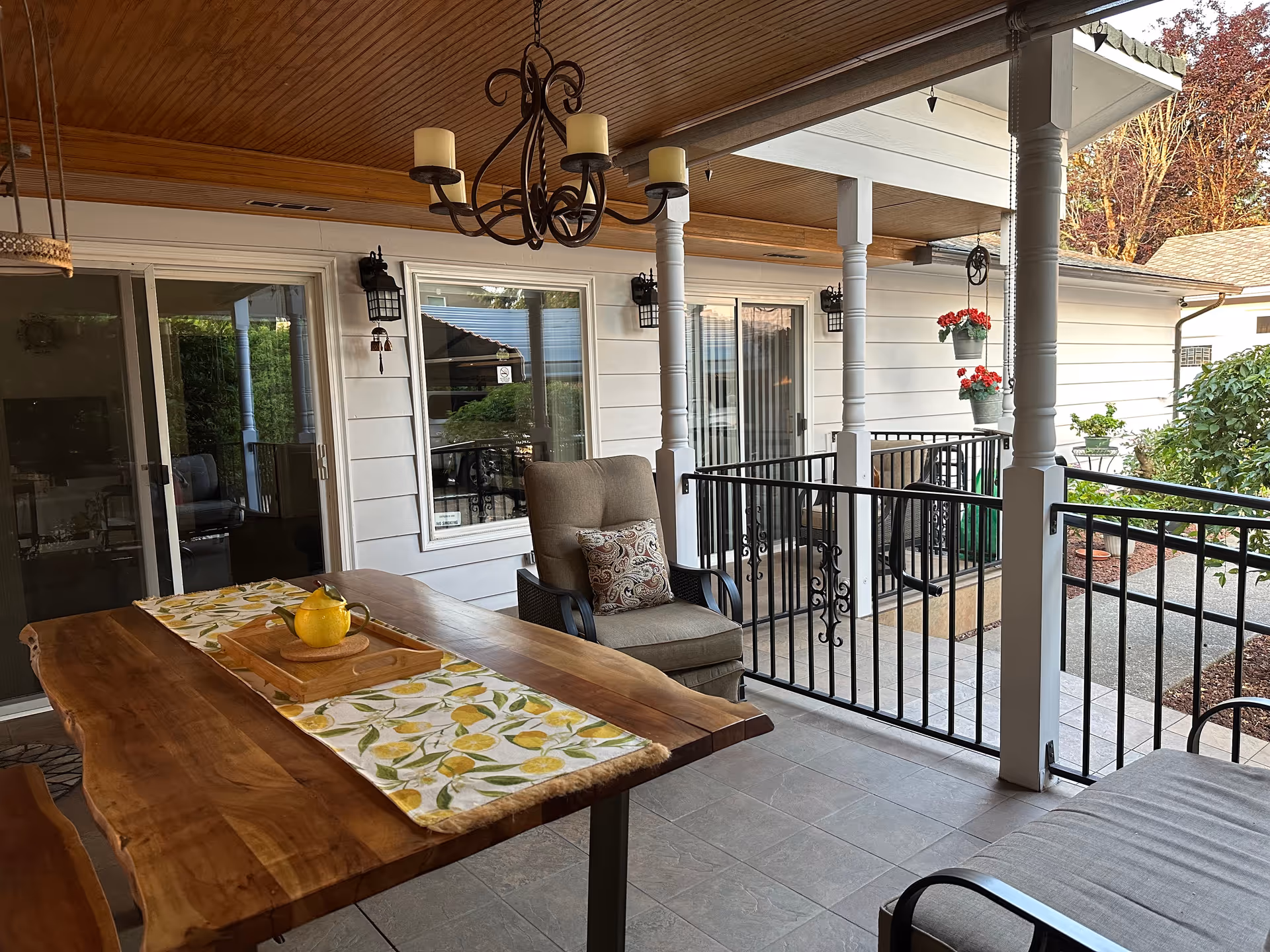 Covered outdoor patio with a wooden table, cushioned chairs, railing, and hanging chandelier.