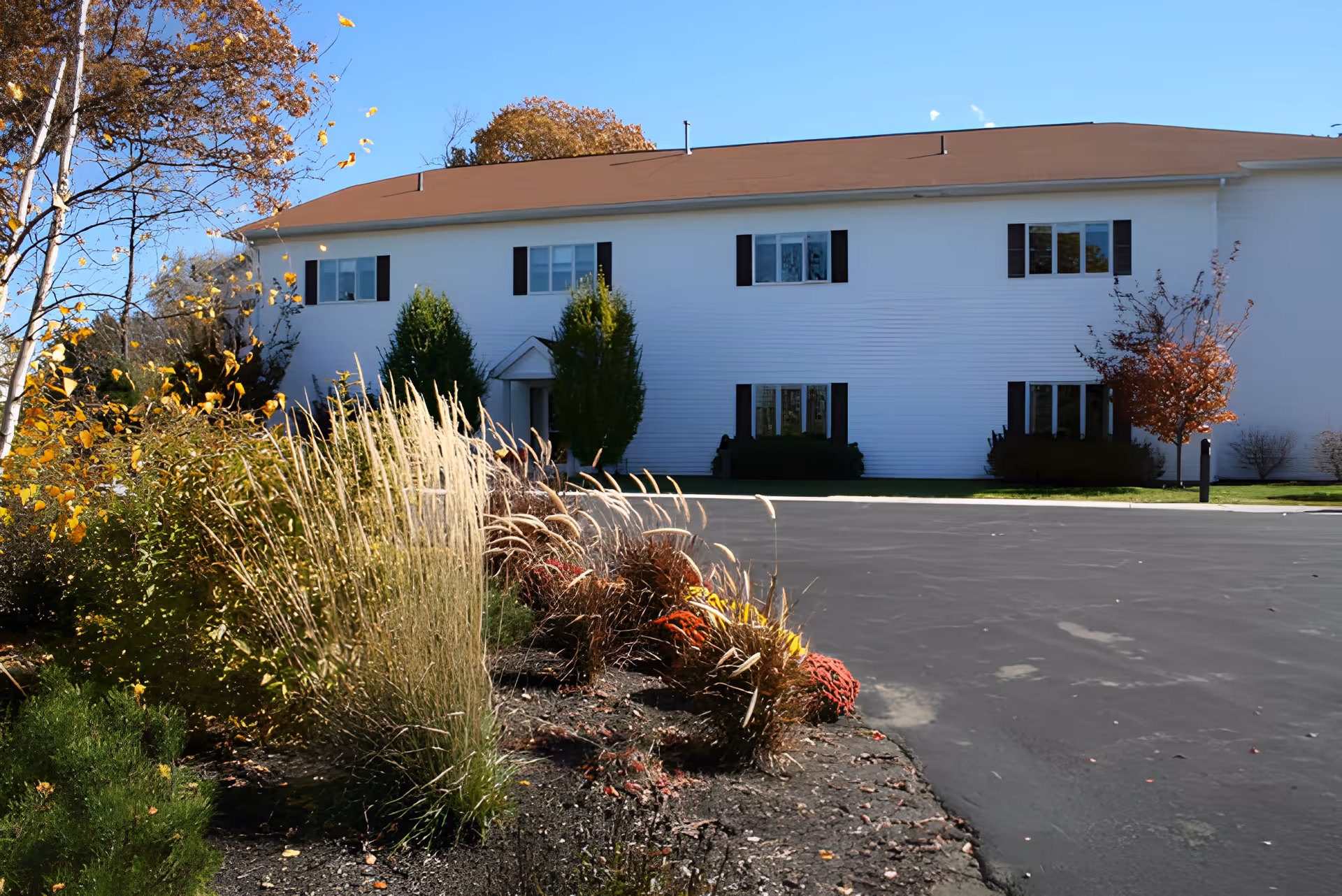 Exterior view of a two-story white building with a brown roof, surrounded by landscaped bushes, ornamental grasses, and small trees with autumn foliage under a clear blue sky.
