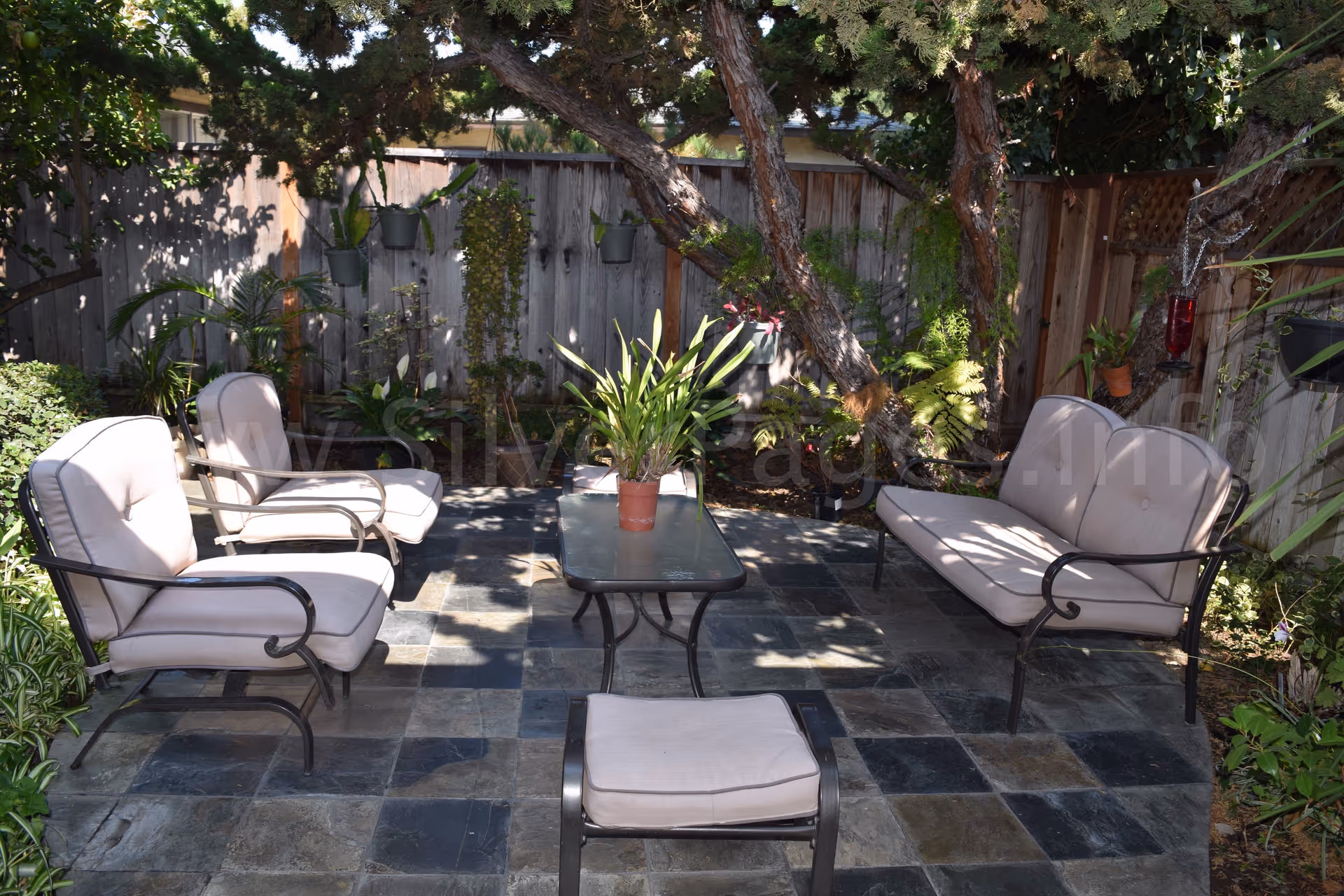 Outdoor patio with cushioned seating, a glass table, and potted plants near a wooden fence.