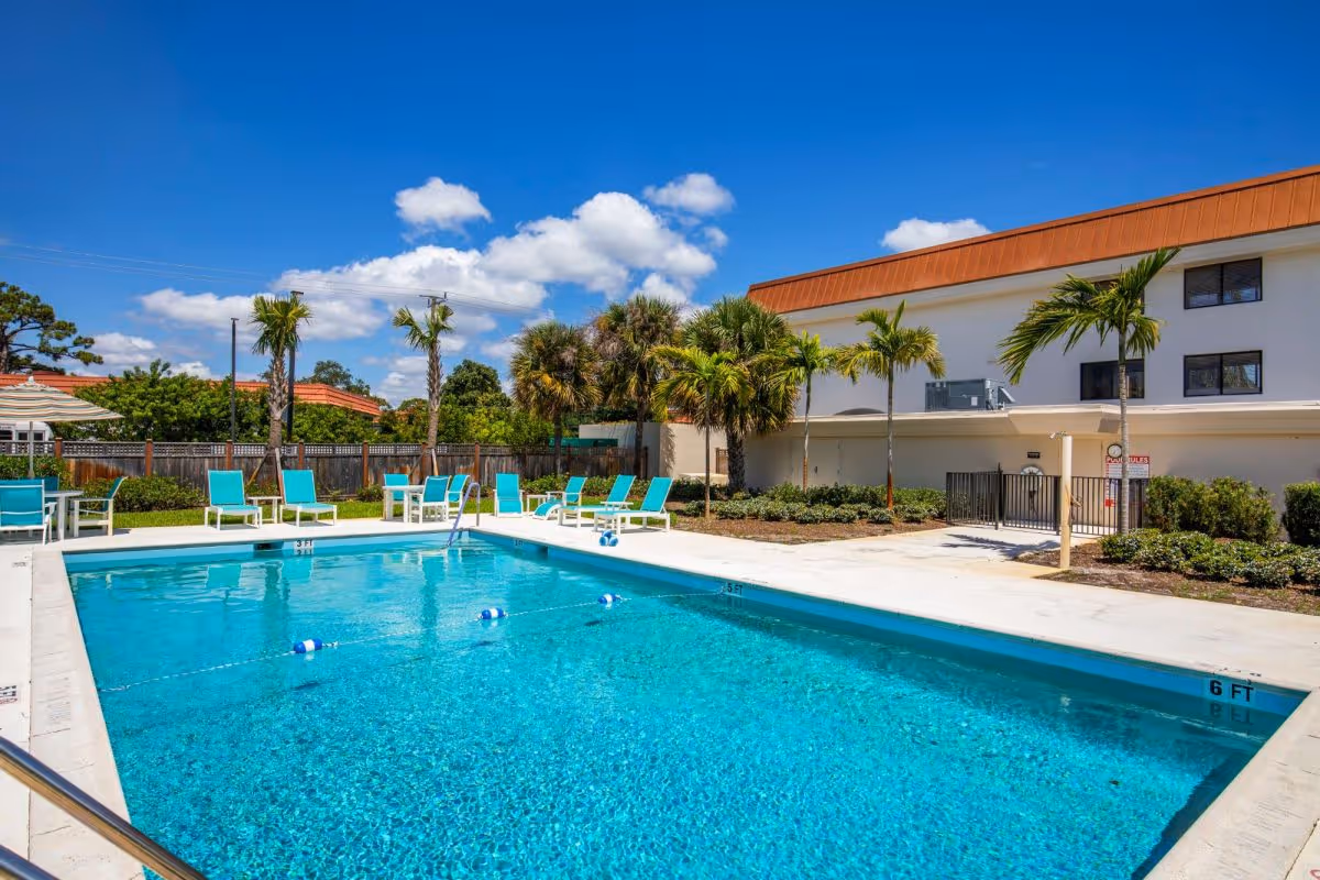 Outdoor swimming pool with clear blue water surrounded by white concrete deck and turquoise lounge chairs. Palm trees and shrubs are planted around the pool area. A two-story building with a brown roof is visible in the background under a bright blue sky with scattered white clouds.