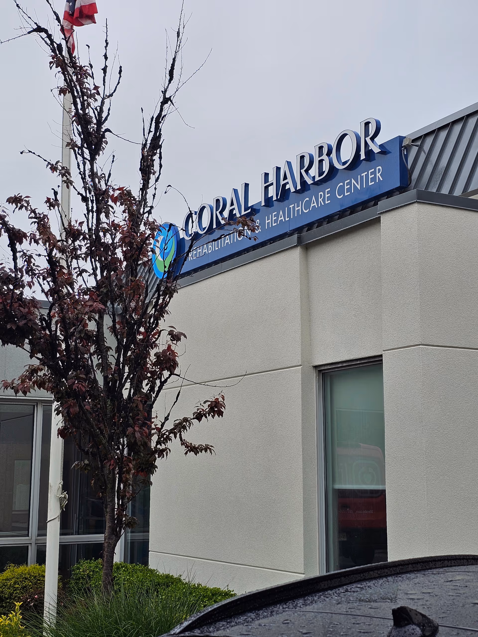 Exterior of Coral Harbor Rehabilitation and Healthcare Center with a blue sign and tree in foreground.