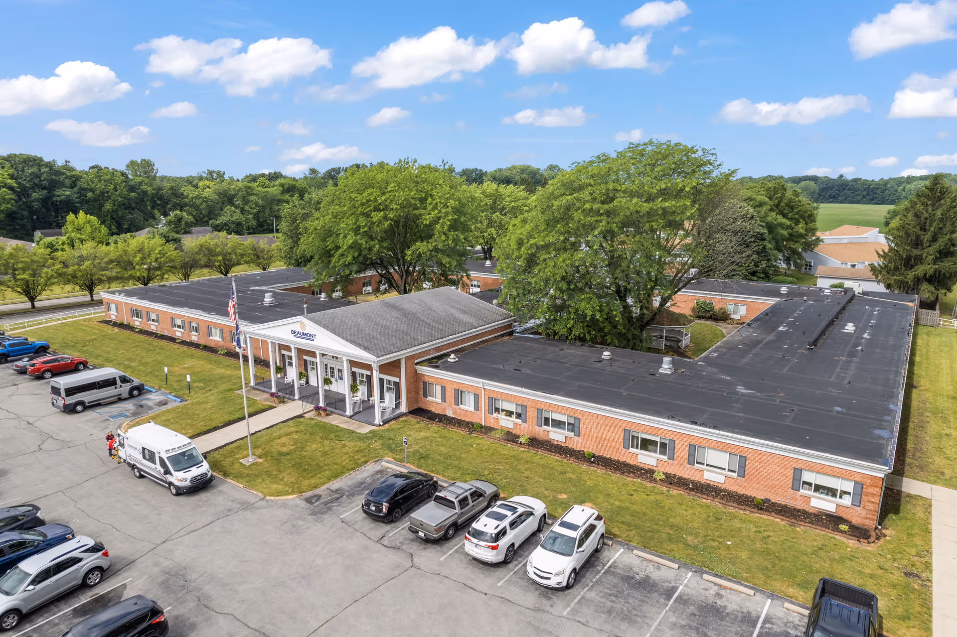 Aerial view of a single-story brick senior living facility with a front entrance, parking lot, and surrounding trees.