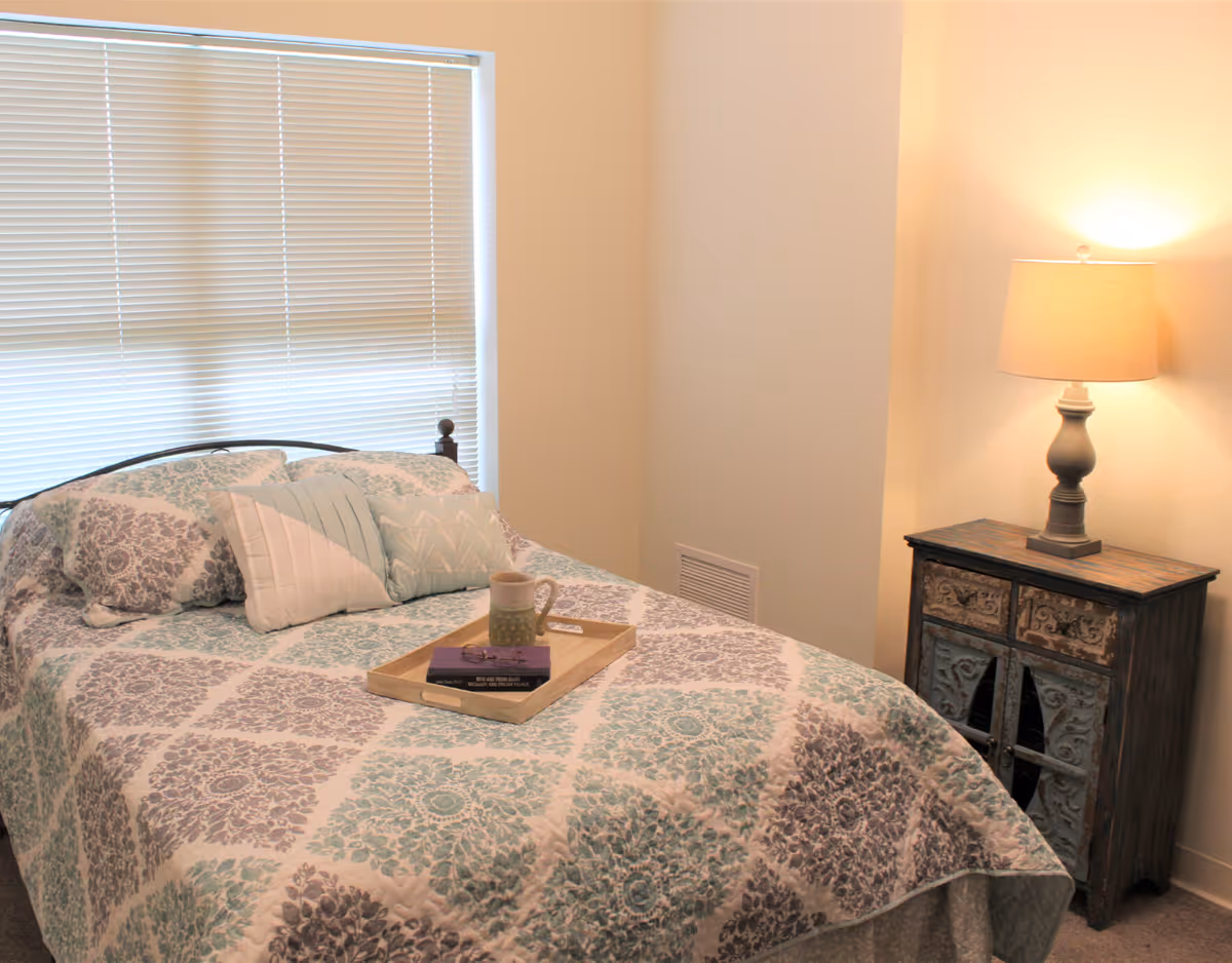Small bedroom with a patterned bed and pillows, a tray with a mug on the bed, and a lit lamp on a carved wooden nightstand.