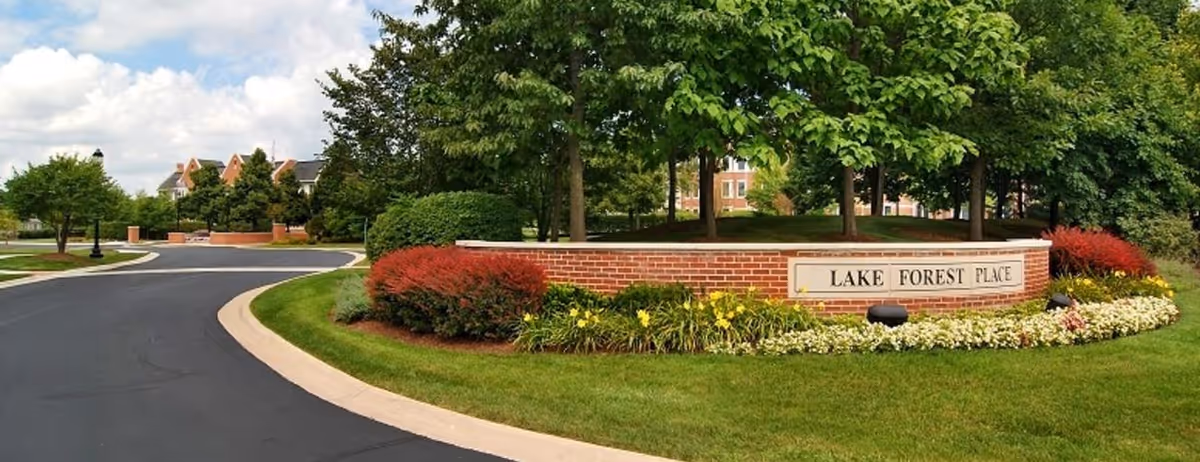 Curved driveway leads to a brick sign reading Lake Forest Place, surrounded by lush landscaping.