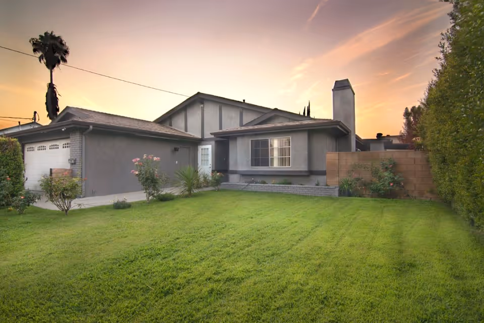 Front exterior of a single-story house with a green lawn, driveway and garage under a sunset sky.