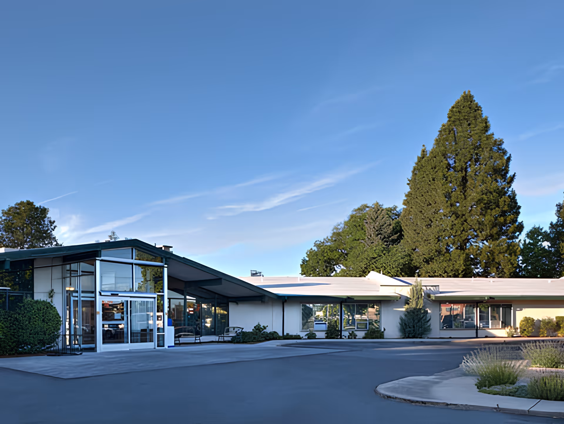 Exterior view of a single-story building with large windows and a glass entrance door, surrounded by greenery and tall trees under a clear blue sky.