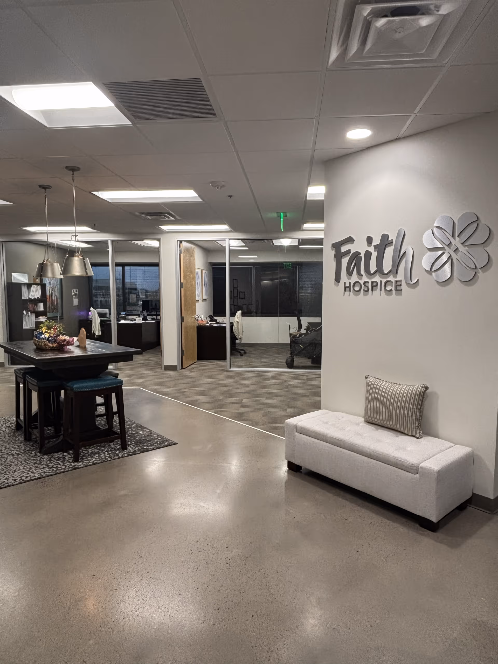 Lobby-like interior with a bench, high table, and Faith Hospice signage on the wall.