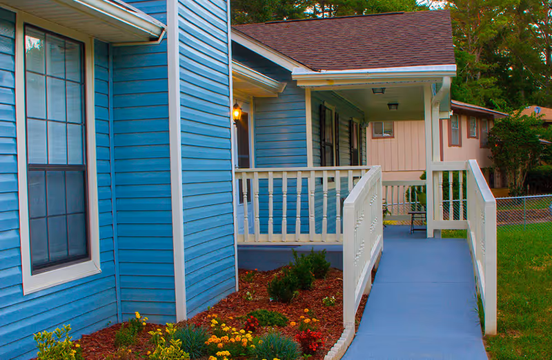Blue residential building exterior with a ramp, white railing, and landscaped garden.