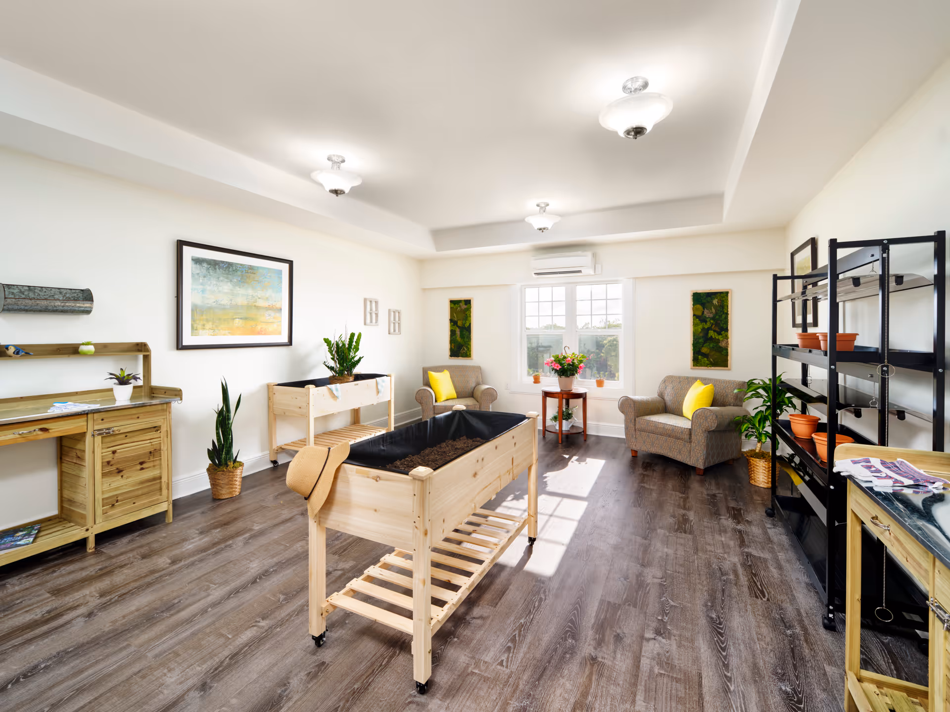 Bright senior living common room with sofas, chairs, plants, shelving, and a central wooden planter.