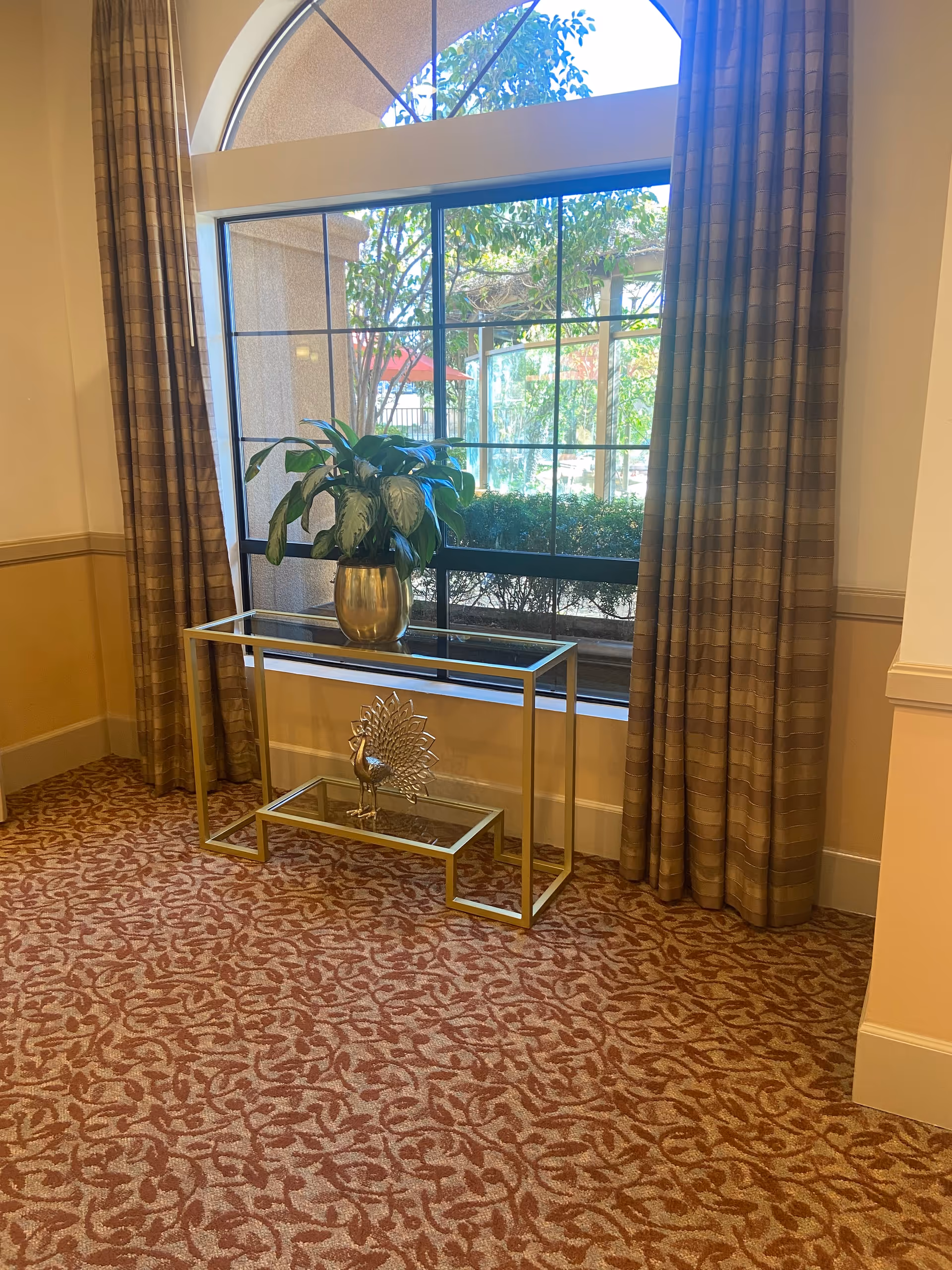 Gold-framed console table with a plant beside a large window and patterned carpet.