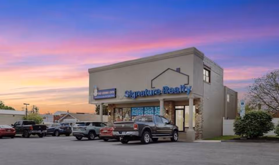 Exterior storefront building with a Signature Realty sign at sunset and parked cars.
