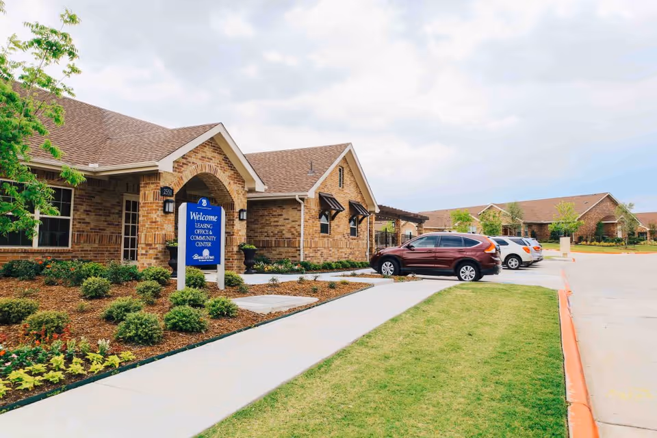 Exterior view of a brick building with a sign that reads 'Welcome Leasing Office & Community Center' at Emerald Cottages of Stonebridge. There are several parked cars along the driveway and landscaped greenery around the building.