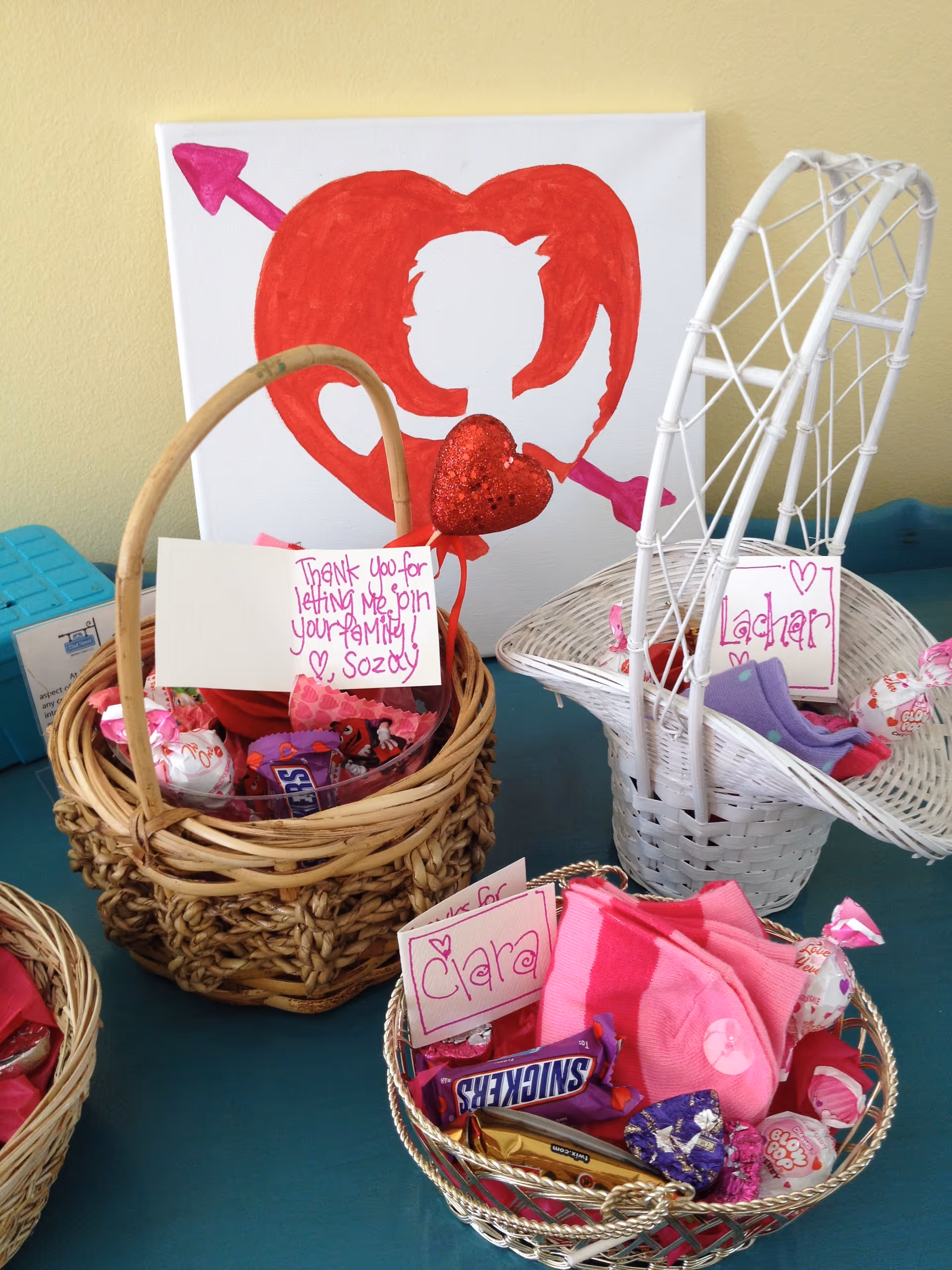 Three baskets filled with candy and small gifts, each with a handwritten note. In the background, there is a canvas with a red heart and a white silhouette of a person with an arrow through the heart. The baskets are placed on a blue surface.