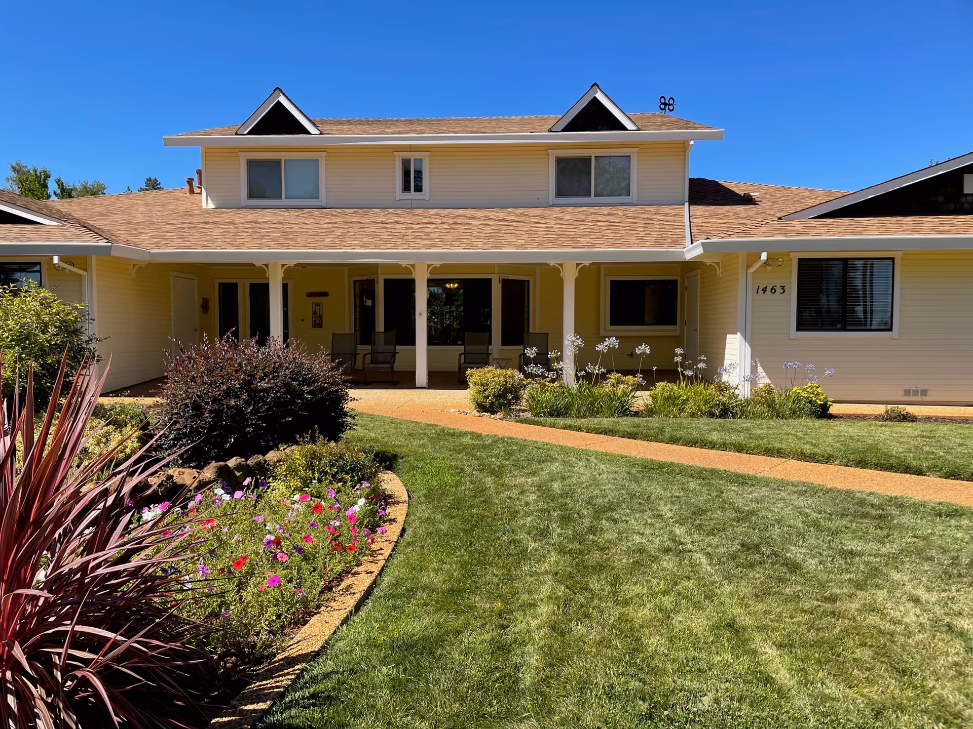 Front exterior of a two-story light-yellow cottage with a covered porch, lawn, and flower beds.