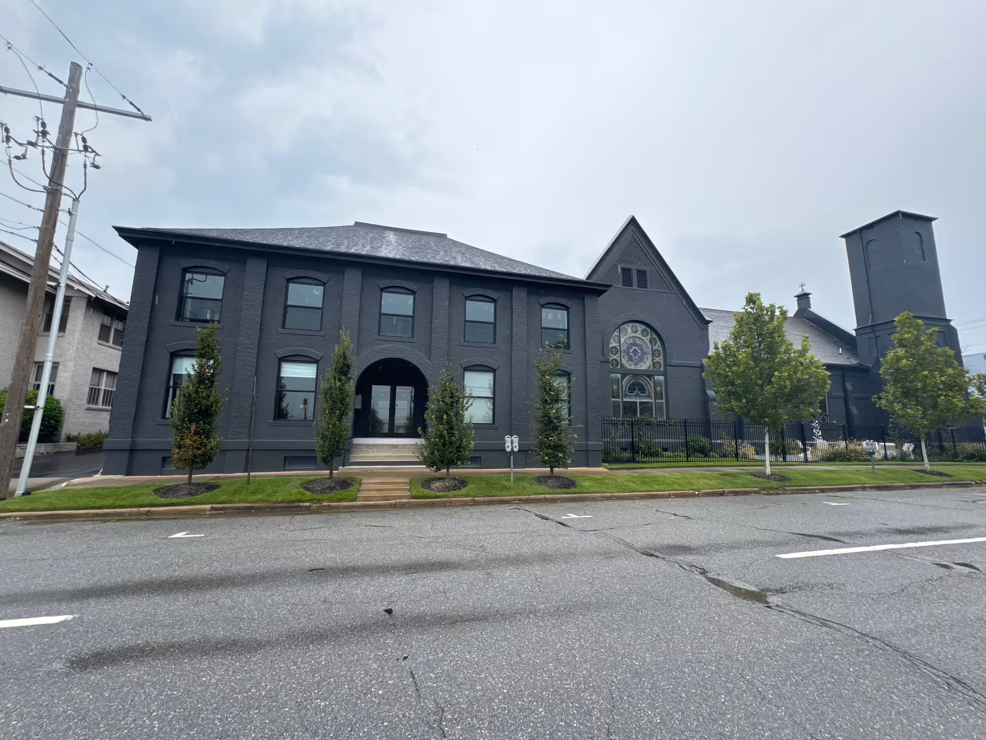 Dark gray brick building with arched windows, small trees, and a front entrance along a street.