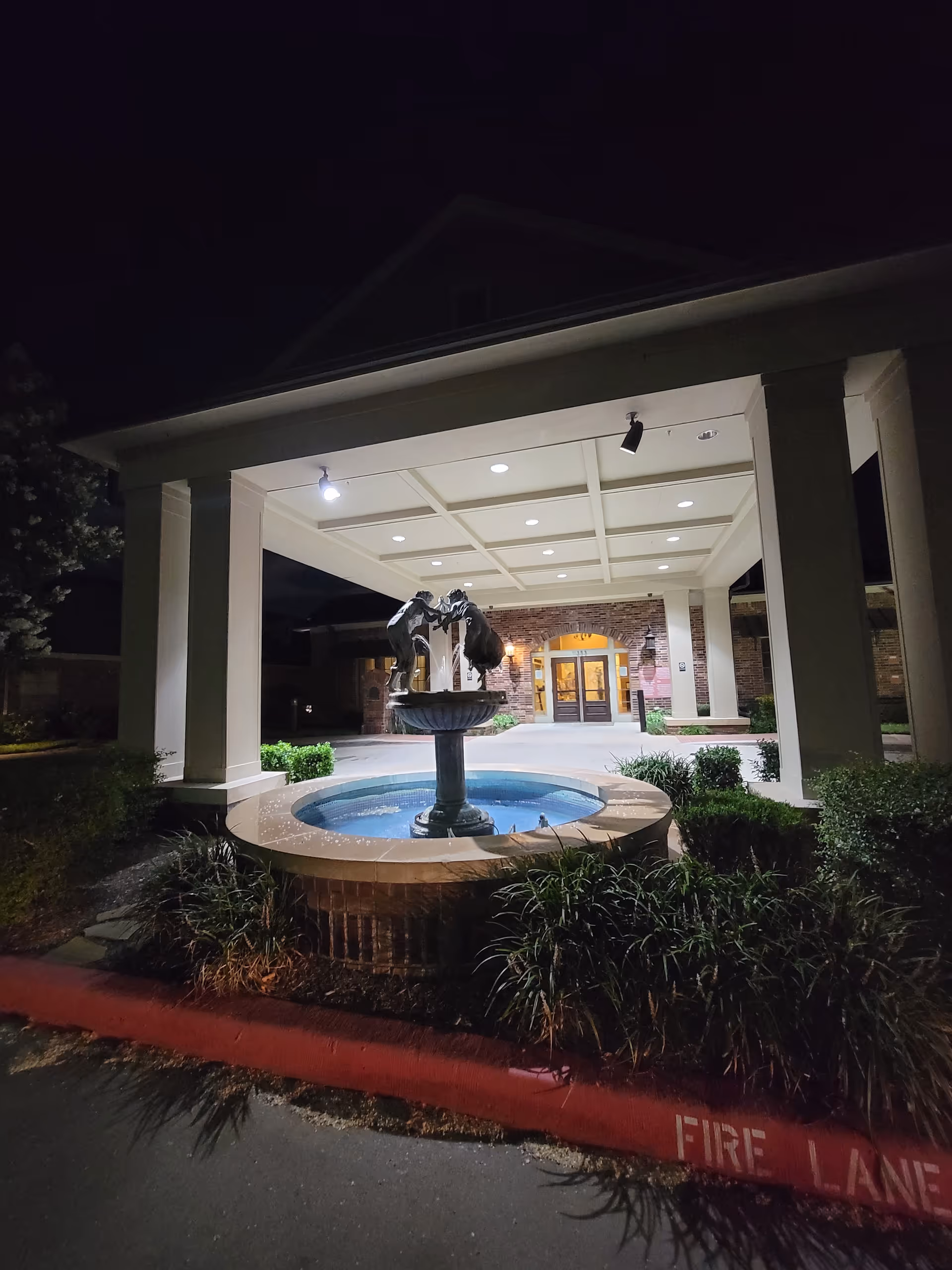 Night exterior of The Crescent facility with a lit entry, fountain, columns, and landscaping.