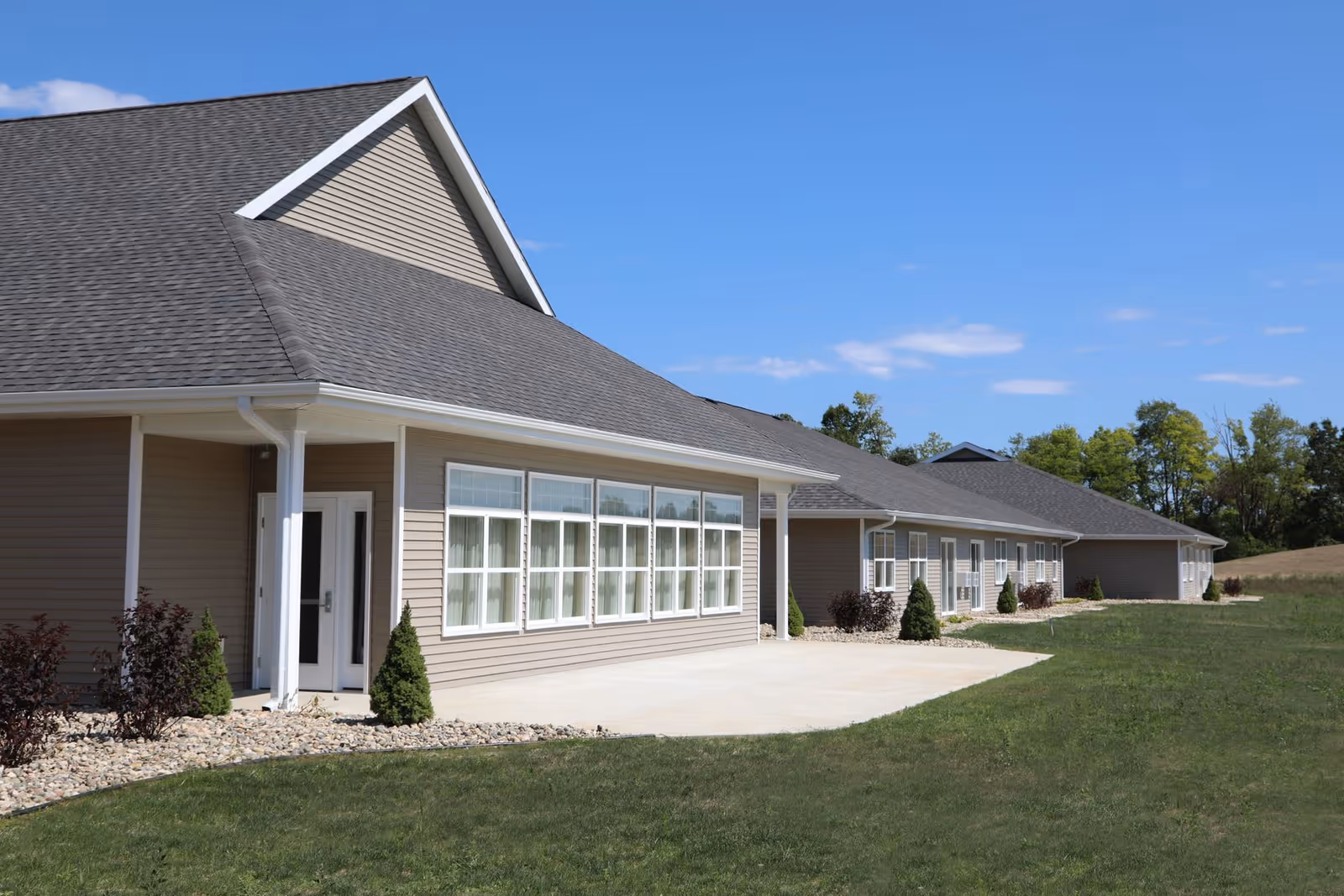 Exterior view of a single-story building with beige siding and a gray shingled roof under a clear blue sky. The building has multiple large windows with white frames and a concrete walkway surrounded by grass and small shrubs.