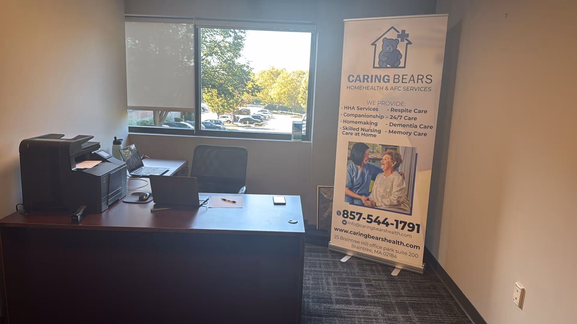 Interior office with desk, printer, laptop, and a Caring Bears banner near window.
