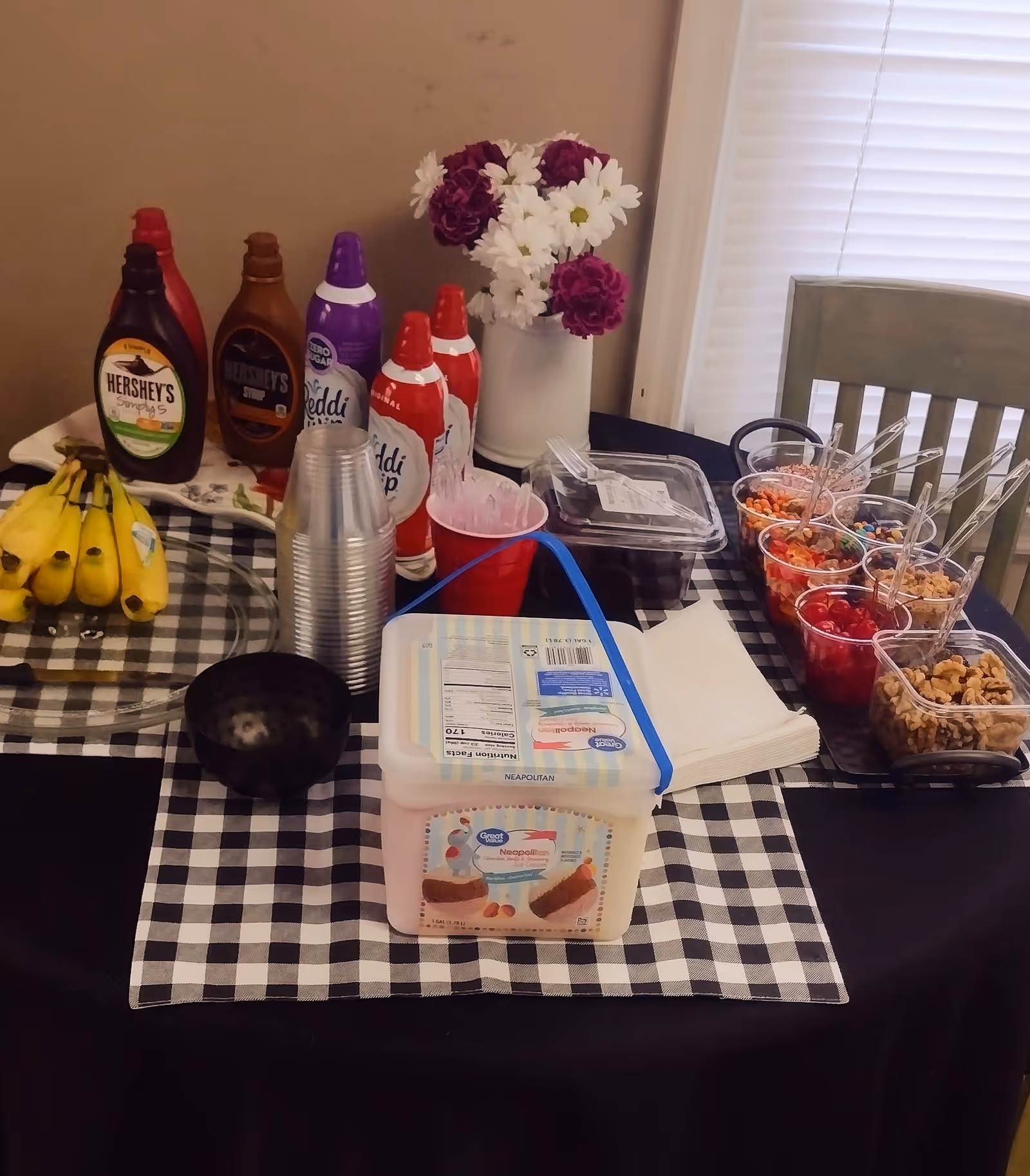 A checkered dining table with sauces, snacks, cups, and a bouquet of flowers.