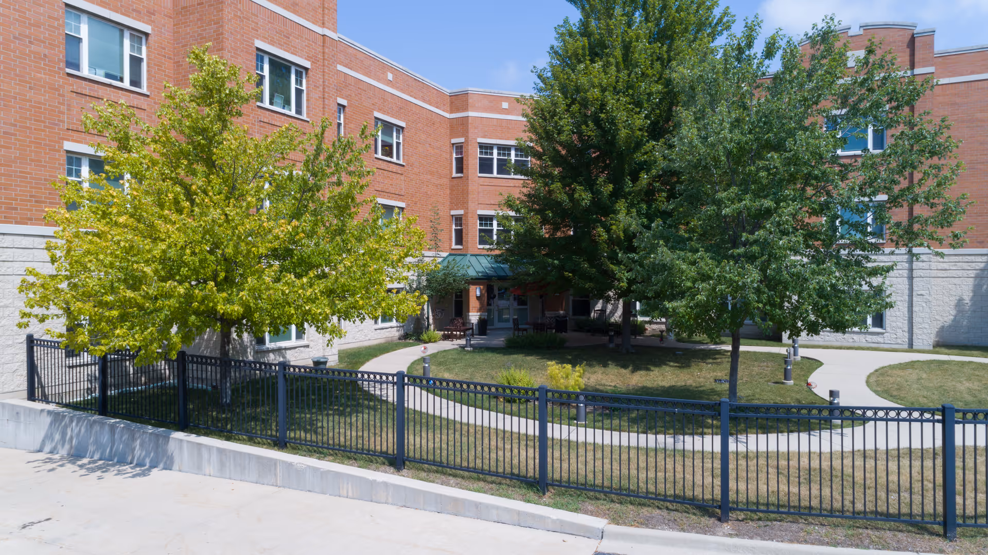Red brick senior living building with a fenced front courtyard, trees, and a curved walkway to the entrance.