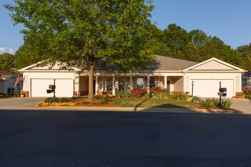Single-story building front with a central porch flanked by two garages, a large tree, mailboxes and landscaped flower beds.