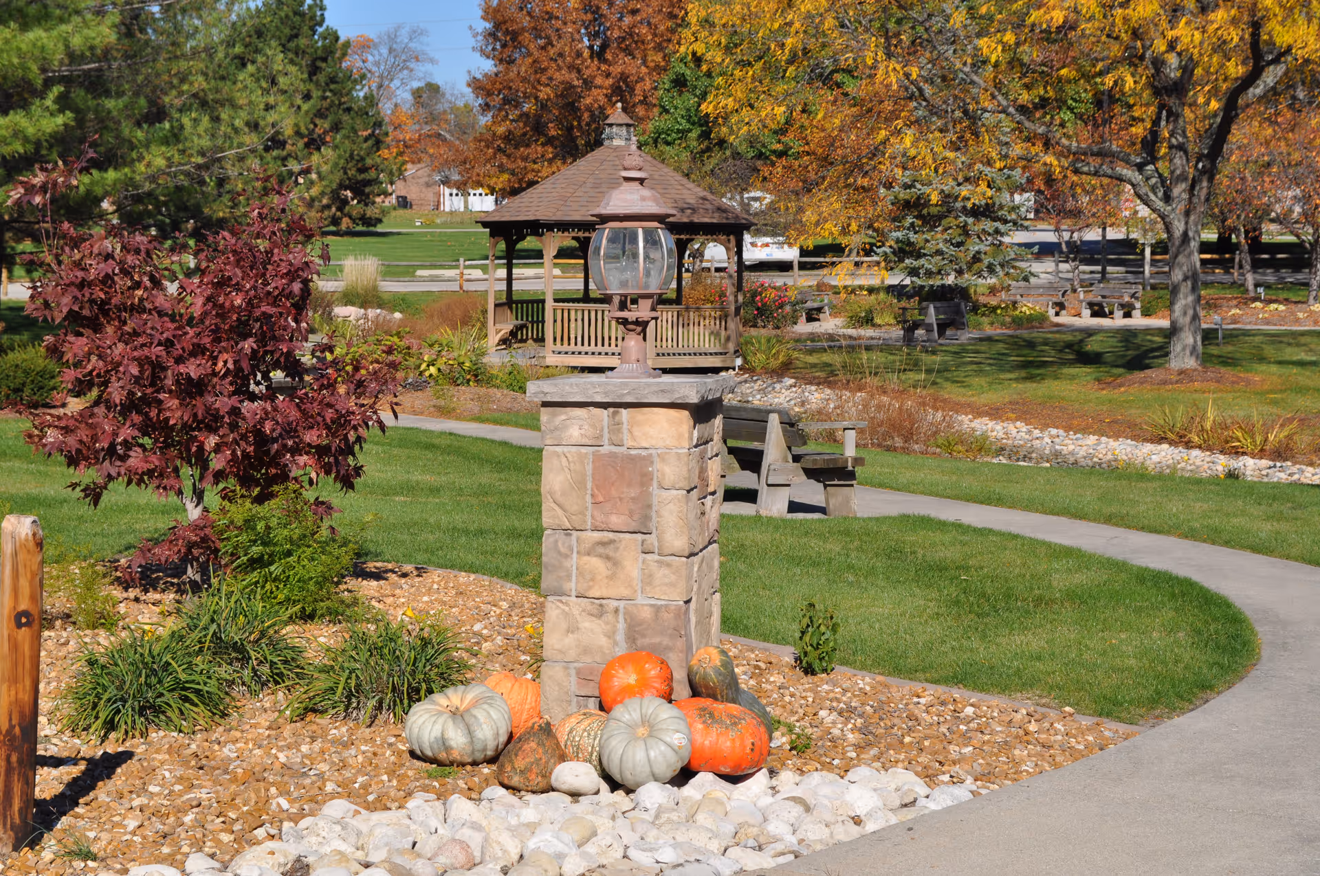 A landscaped outdoor area at Westminster Village featuring a stone pillar with a lantern on top, surrounded by various pumpkins and gourds. There is a curved concrete pathway, green grass, trees with autumn foliage, wooden benches, and a gazebo in the background.