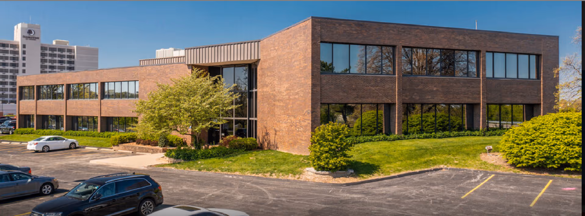 Two-story brick building with large windows, parking lot, and landscaped surroundings.