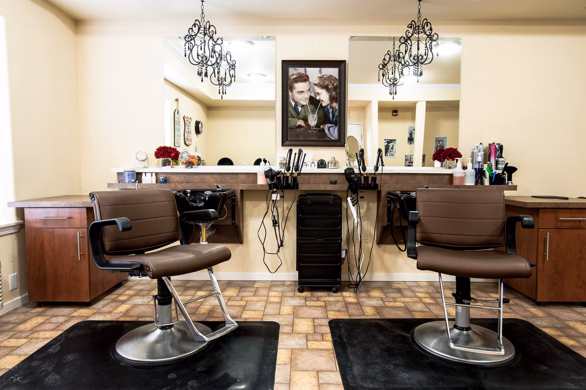 Two salon styling chairs and workstations with mirrors, hair tools, and chandeliers in a salon area.