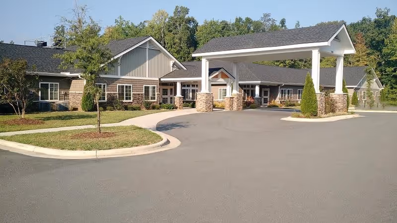 Exterior view of a single-story senior living facility building with a covered entrance supported by stone pillars, surrounded by a paved driveway and landscaped greenery including small trees and shrubs.