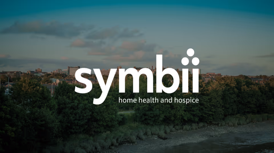 Outdoor riverfront scene with trees and city skyline, overlaid by the Symbii logo.