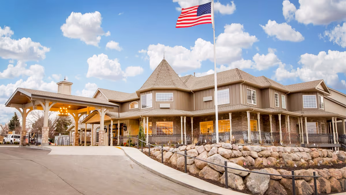 Front exterior of a two-story assisted living building with a covered entrance, American flag, and rock landscaping under a blue sky.