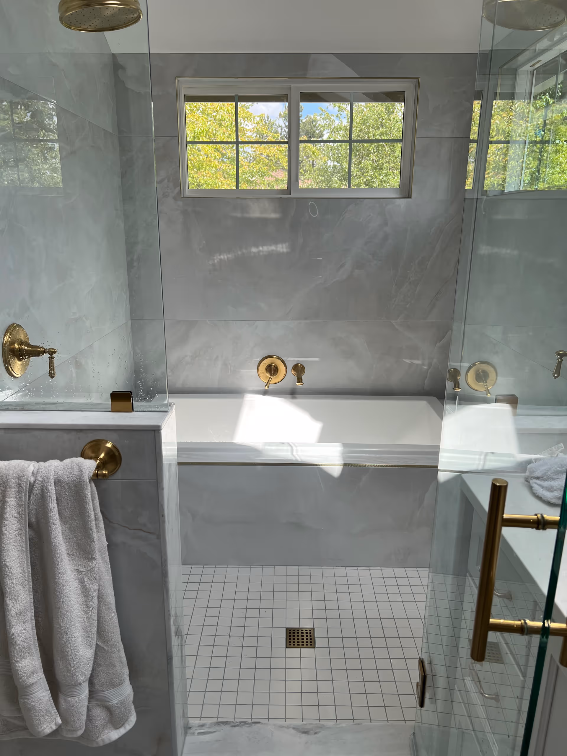 Marble-tiled bathroom with glass shower, gold fixtures, towel, and a small window.