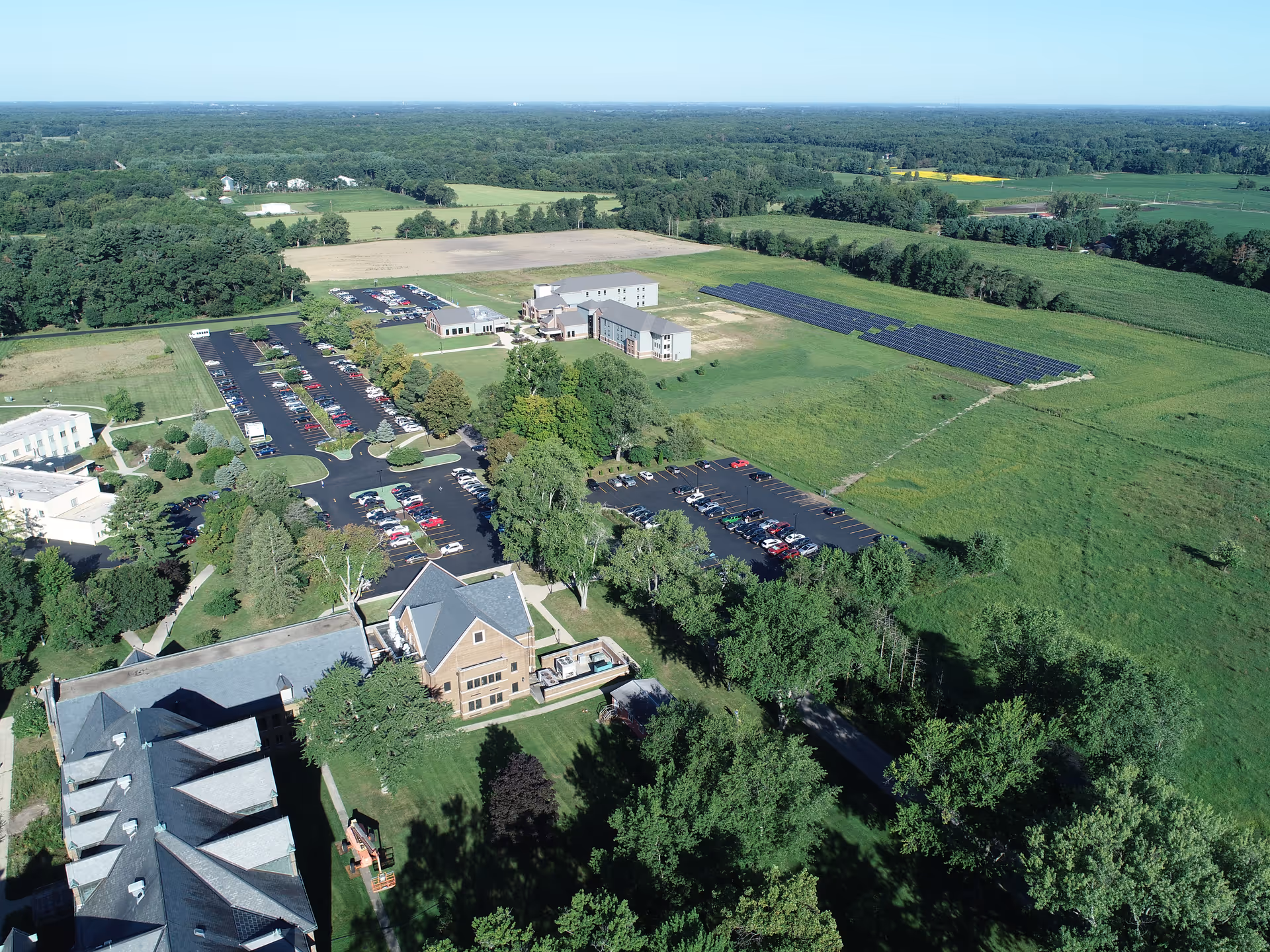 Aerial view of the Catherine Kasper Life Center facility showing multiple buildings, parking lots with cars, green lawns, trees, and a large field with solar panels. Surrounding the facility are expansive green fields and wooded areas under a clear blue sky.