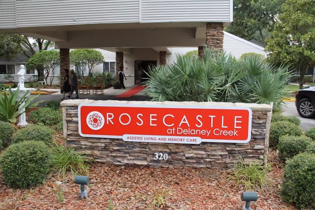 Stone sign reading "Rosecastle at Delaney Creek Assisted Living and Memory Care" in front of the facility entrance with landscaping and a covered drop-off.