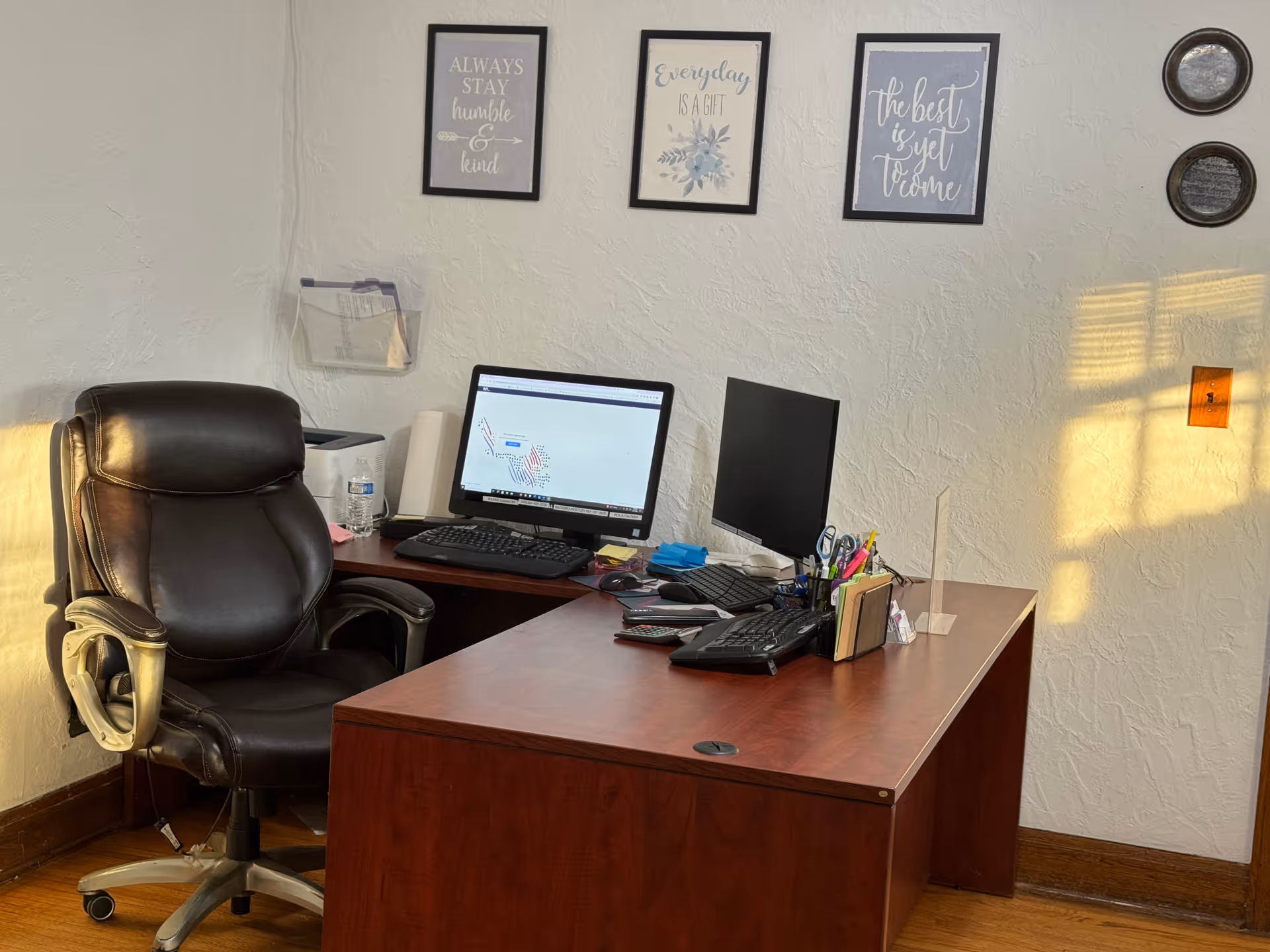 A dark wooden desk with two monitors, office supplies, and a black leather chair.