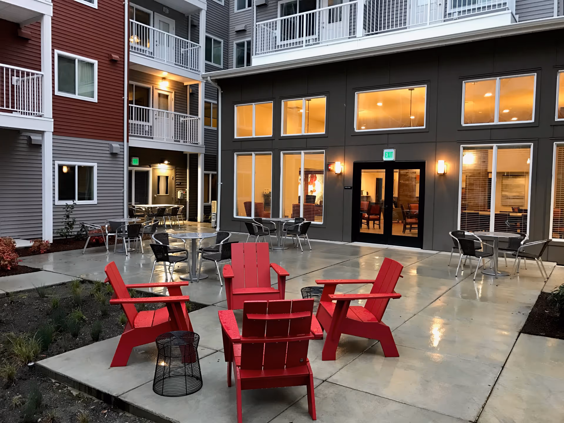 Outdoor courtyard area of a senior living facility with four red Adirondack chairs arranged around a small black metal table on a concrete patio. Several metal tables with chairs are placed around the patio. The building exterior features large windows and doors with warm interior lighting visible inside. The building has multiple floors with balconies and a mix of red and gray siding.