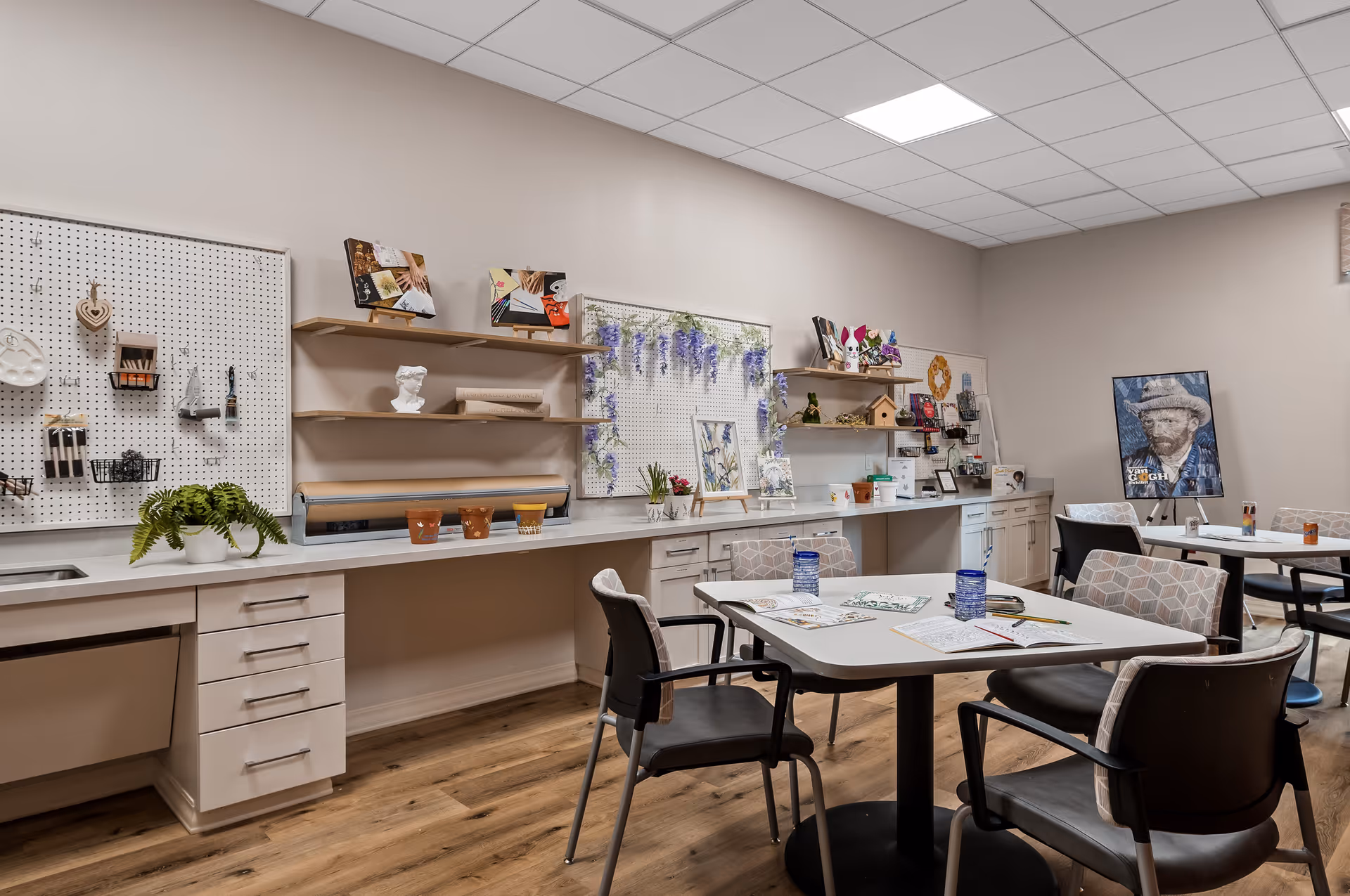 A well-lit activity room with tables and chairs arranged for group activities. The tables have coloring books, pencils, and cups on them. The back wall features pegboards with various art supplies, small decorative items, and shelves displaying craft projects and plants. A framed picture of Vincent van Gogh is displayed on an easel in the corner.