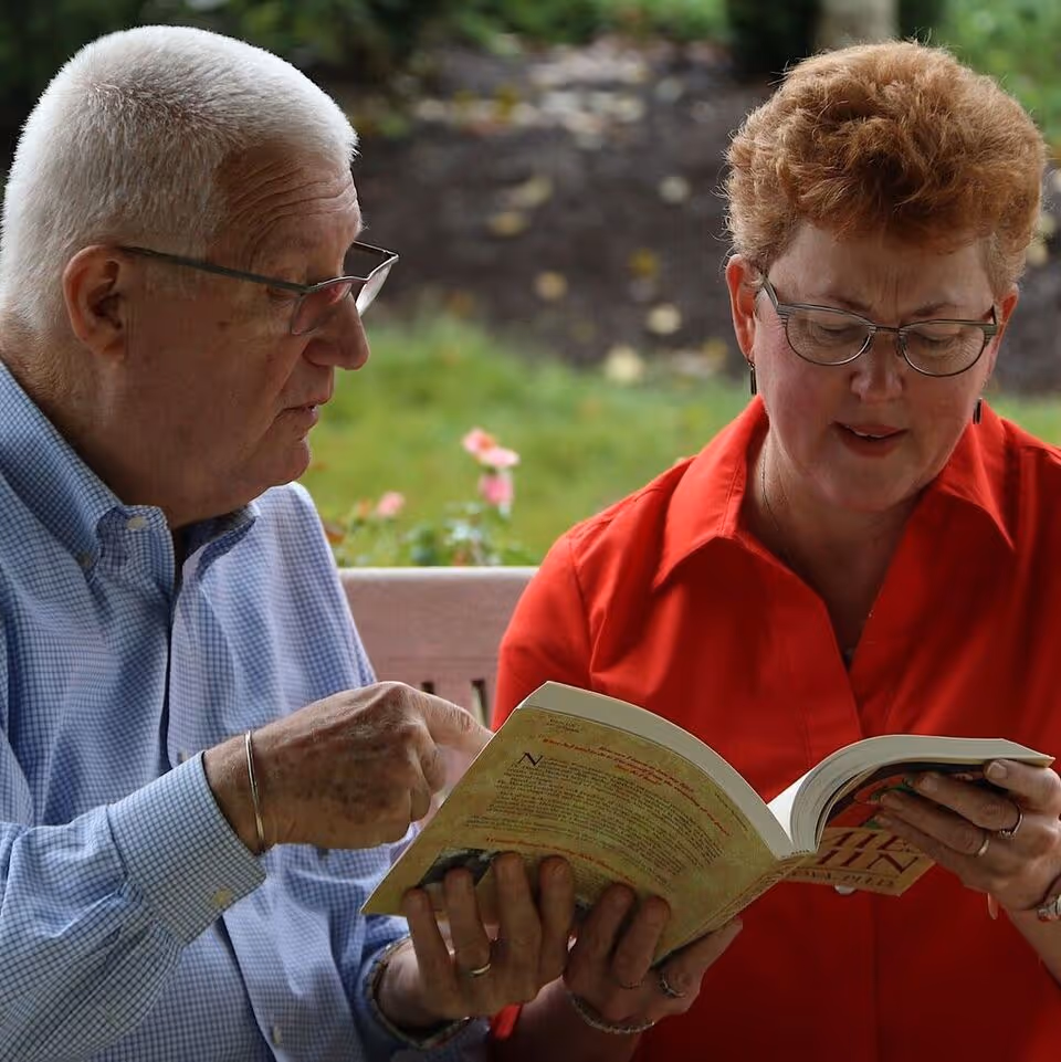 Two seniors sit on a bench outdoors, reading a book together.