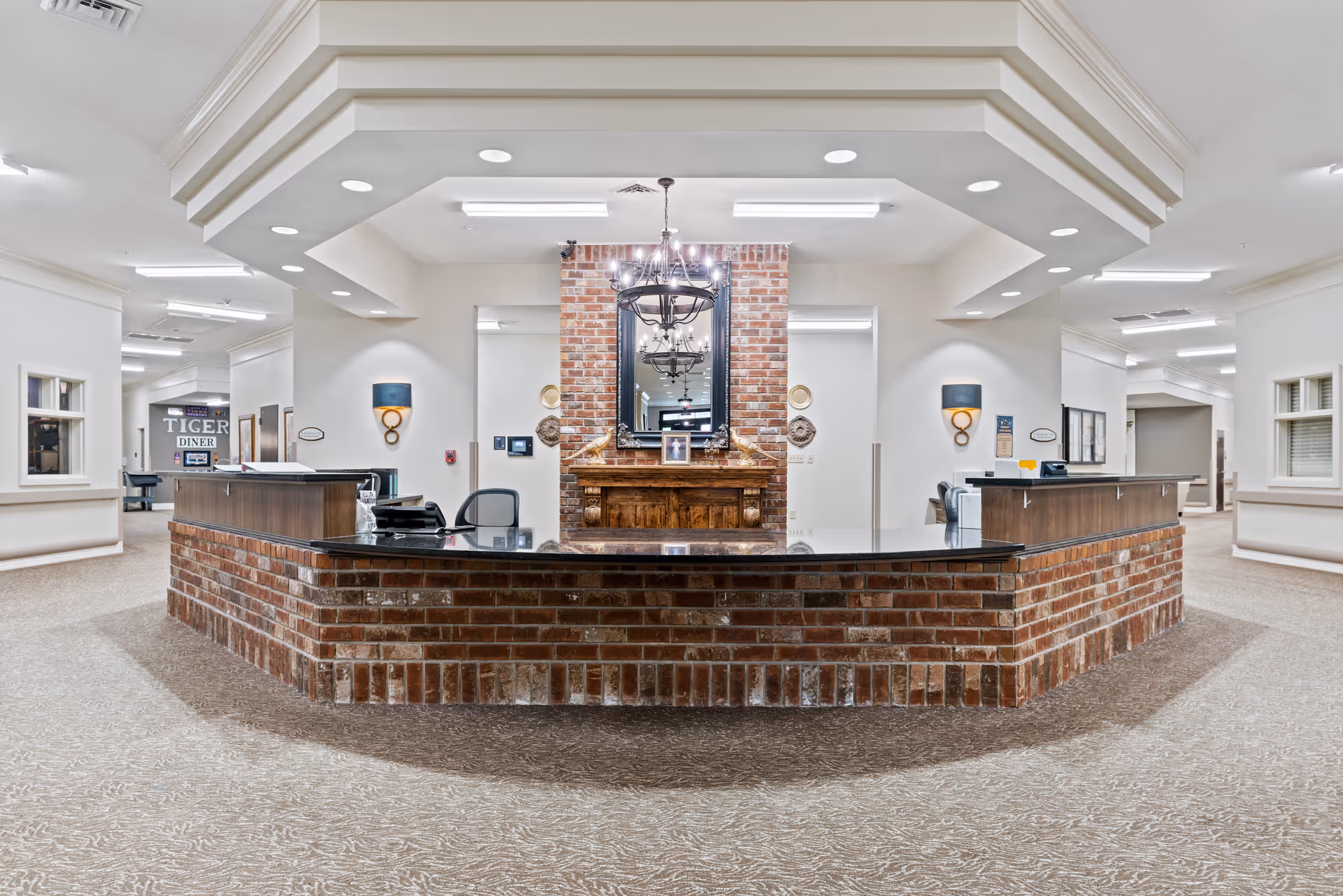 A spacious reception area with a large brick front desk and black countertop. Behind the desk is a decorative brick wall with a wooden mantel, a large mirror, and a chandelier hanging from the ceiling. The area is well-lit with recessed lighting and wall sconces. The carpeted floor and neutral-colored walls create a welcoming atmosphere. In the background, a sign reading 'Tiger Diner' is visible down a hallway.