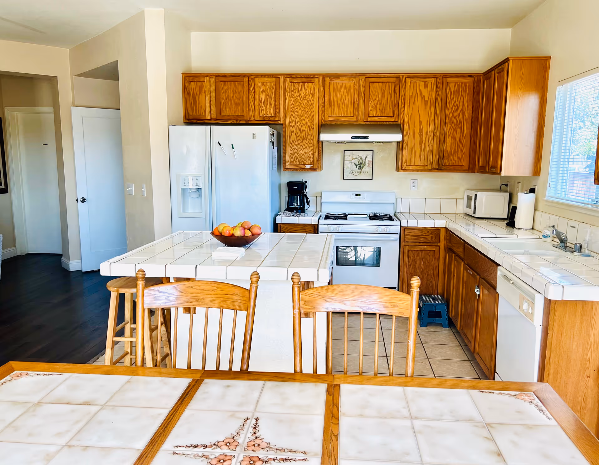 Bright kitchen with wooden cabinets, white tiled countertops, a white refrigerator, stove, microwave, coffee maker, and a bowl of apples on the kitchen island. Wooden chairs are visible in the foreground and a window lets in natural light on the right.