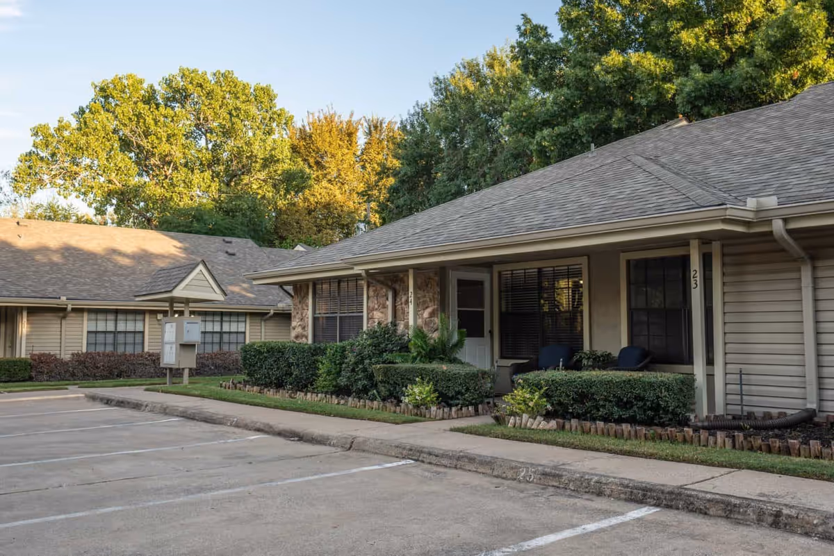Exterior view of a single-story residential building with a porch, shrubs, and a parking area in front. The building has beige siding, a shingled roof, and large windows with blinds. Trees with green and yellow leaves are visible in the background under a clear sky.