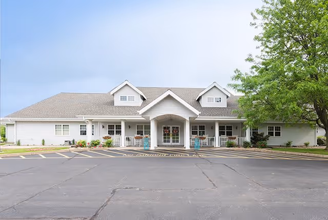 Front exterior view of a single-story senior living facility building with a covered entrance, white siding, multiple windows, a porch with chairs, and a large tree on the right side. The parking lot is in front with marked no parking zones.