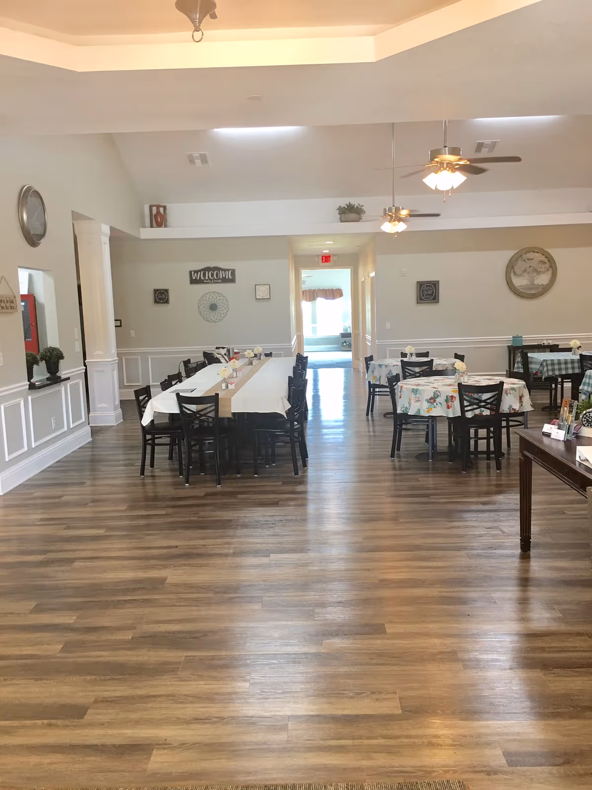 A spacious dining room in an assisted living facility with wooden flooring, several tables covered with white and floral tablecloths, and black chairs arranged around them. The room features ceiling fans with lights, wall decorations including a 'Welcome' sign, and a hallway leading to another room with natural light coming through the windows.