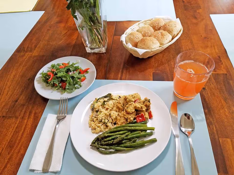 A wooden dining table set with a meal including a plate of rice, cooked asparagus, and a vegetable dish, a small side salad on a separate plate, a basket of bread rolls, and a glass of orange-colored beverage. Silverware and a napkin are placed on a light blue placemat, with a vase of flowers in the background.